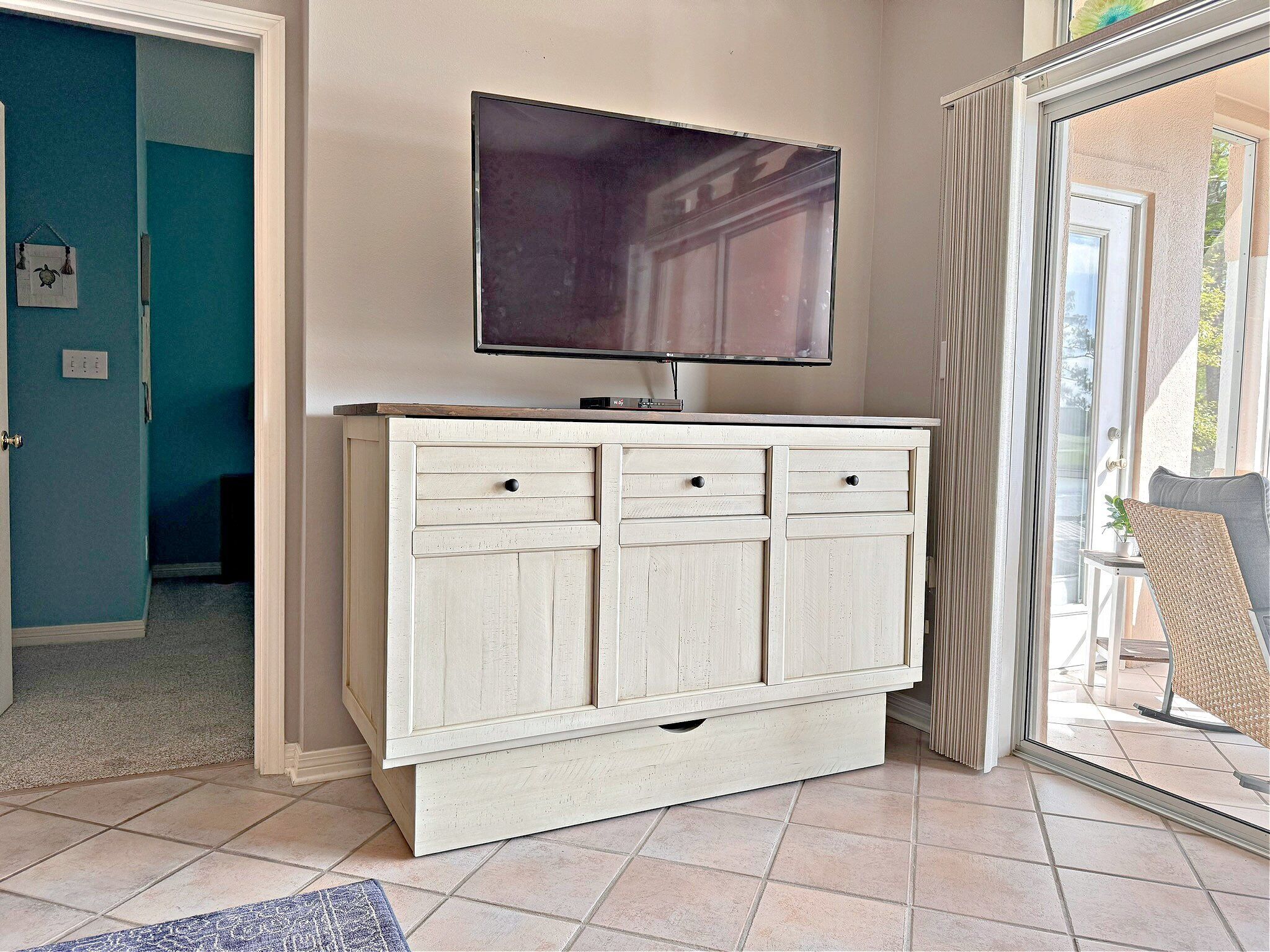 A room with a white cabinet, a flat-screen TV mounted above it, and a sliding glass door leading outside.