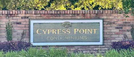 A sign for Cypress Point Condominiums with a brick wall and greenery in the background.