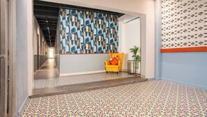 A hallway with patterned floor tiles, a yellow armchair with a colorful cushion, and a decorative wall panel. - Fit for a Queen @ Somos Hotel (San Juan)