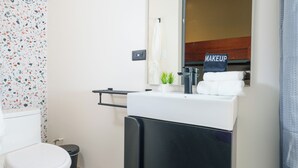 A bathroom with a white toilet, a black trash bin, a sink with a mirror above it, and a towel rack. - Queen Bee @ Somos Hotel (SanJuan)