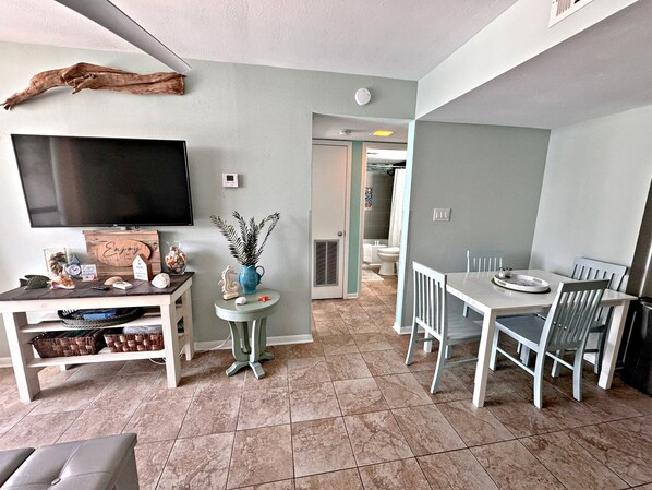 A modern living room with a flat-screen TV, a dining table with chairs, and a small side table with decorative items. - Gulf Shores Surf & Racquet Club (Gulf Shores)