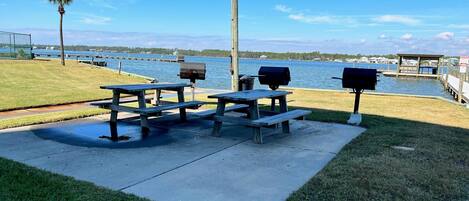 A picnic area with tables and grills by a lake, surrounded by grass and a wooden fence.
