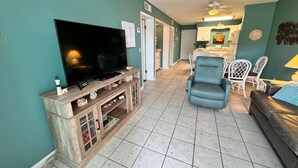 A living room with a TV on a wooden stand, a blue armchair, and a dining area with white chairs. - Gulf Shores Plantation (Gulf Shores)