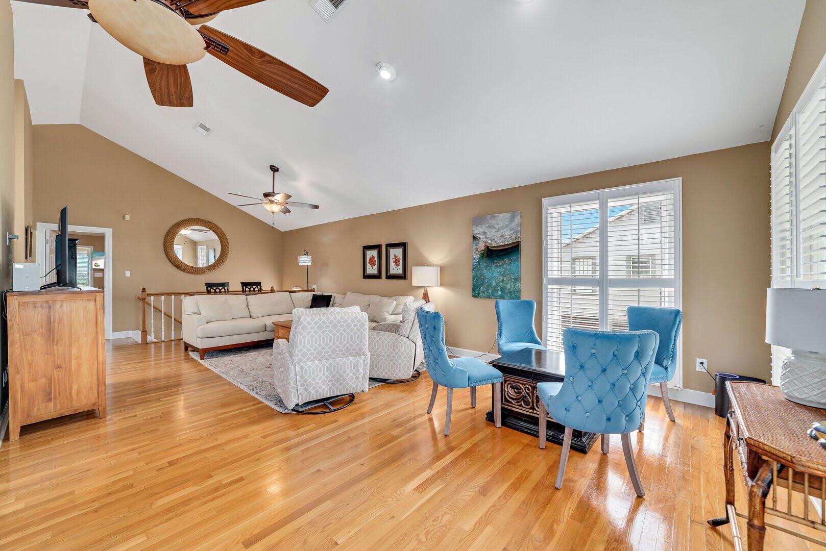 A spacious living room with a wooden floor, a white sofa, blue chairs, a dining table, and a ceiling fan.