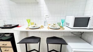 Une cuisine avec des murs recouverts de carreaux blancs, un plan de travail blanc, un four à micro-ondes, deux tabourets et un évier.