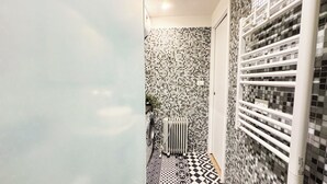 Un couloir étroit, avec un sol et des murs carrelés, un radiateur et une porte blanche.