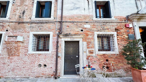Un bâtiment en briques avec une porte noire, portant le numéro 4880, et une enseigne indiquant qu’il s’agit d’une auberge de jeunesse proposant également des chambres privées. Il y a deux fenêtres avec des barreaux en métal, une petite table avec des chaises et un pot de fleurs sur la droite.