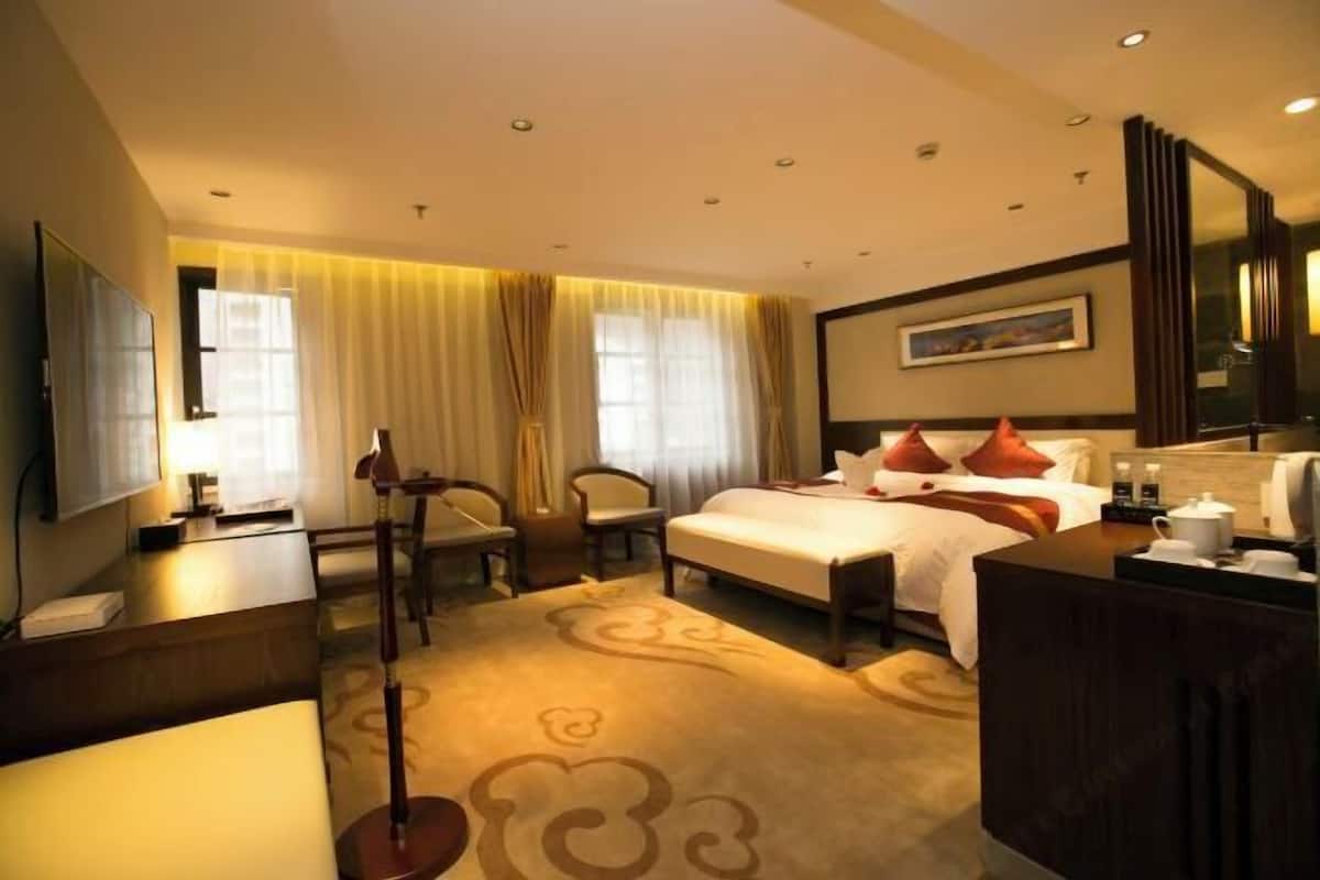 Una habitación de hotel con cama, mesita de noche, lámpara, sillón y un ventanal con cortinas.