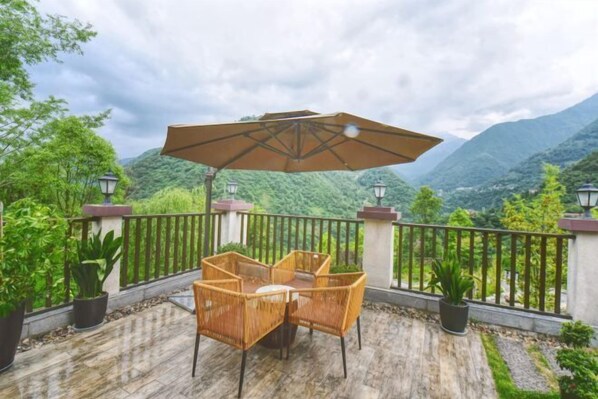 A terrace with wicker furniture and a large umbrella, surrounded by potted plants and a scenic mountain view.