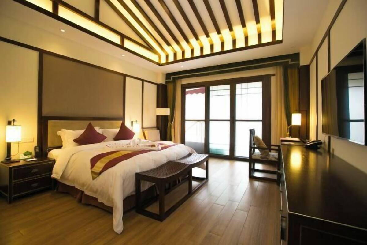 Habitación de hotel con una cama grande, mesitas de noche, un banco, televisión y un amplio ventanal con cortinas.