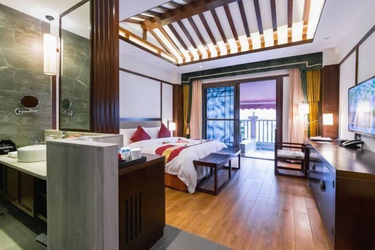 Una habitación de hotel moderna con una cama, mesita de noche y un escritorio con una silla. Hay un ventanal con cortinas y vistas al exterior.