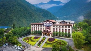 A building with a symmetrical facade, surrounded by lush greenery and mountains. - Gongga cliff Hot Spring Hotel (Luding)