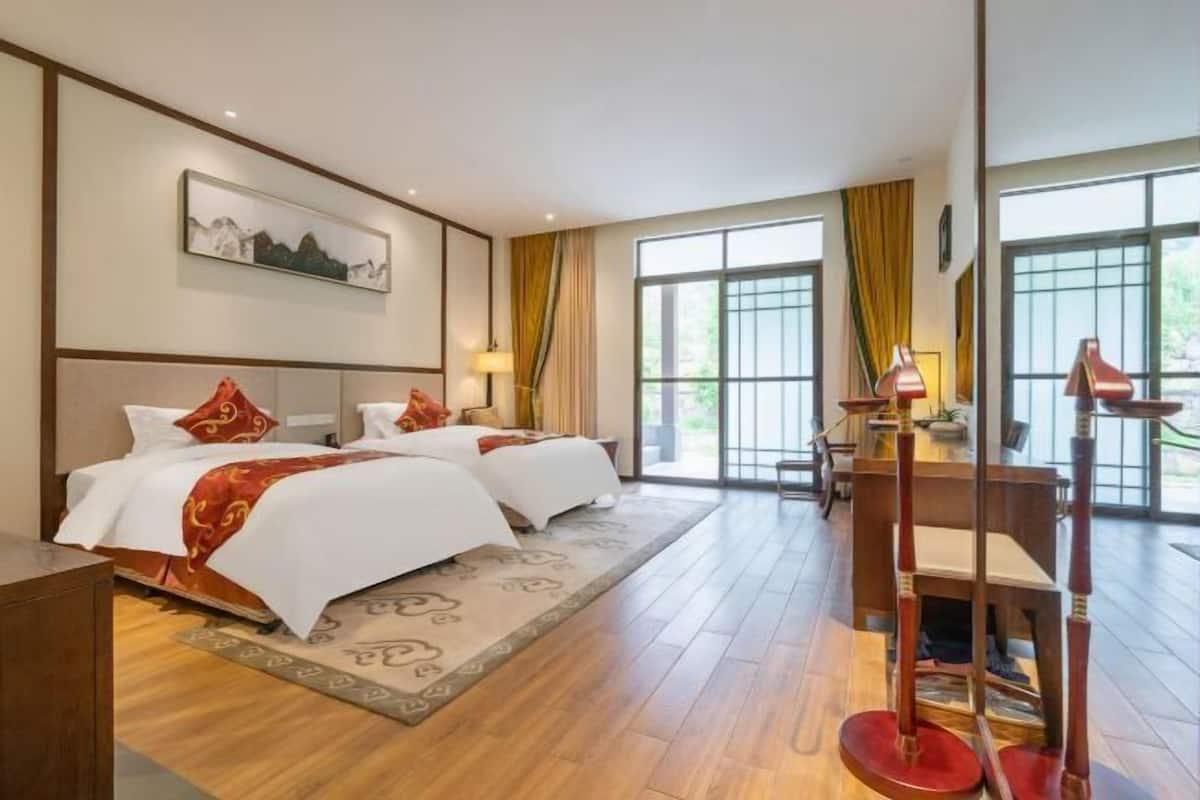 Una habitación de hotel con dos camas, un escritorio, una silla, una lámpara y ventanas amplias.
