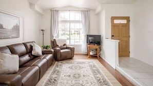 Ein Wohnzimmer mit einem braunen Ledersofa, einem Relaxsessel, einem Fernseher auf einem Schrank, einem großen Fenster mit Vorhängen und einer Holztür.