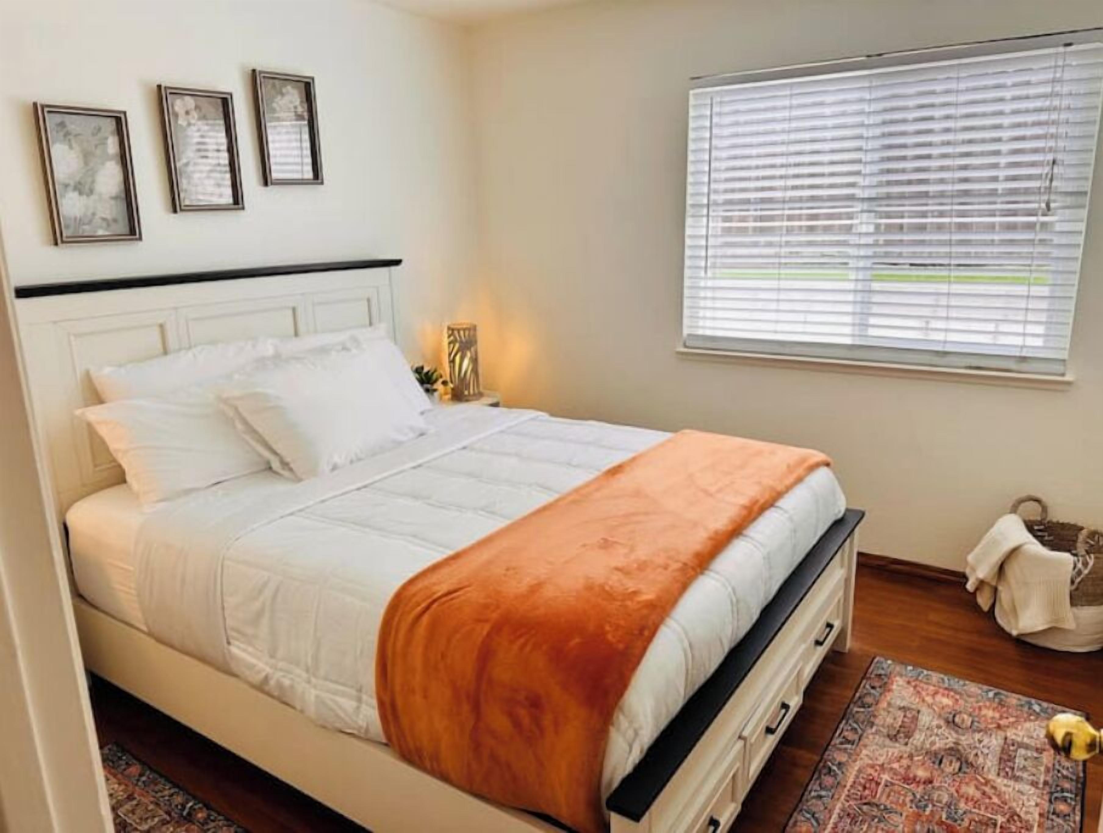A neatly made bed with white bedding and an orange throw blanket, a bedside table with a lamp, and a window with blinds.