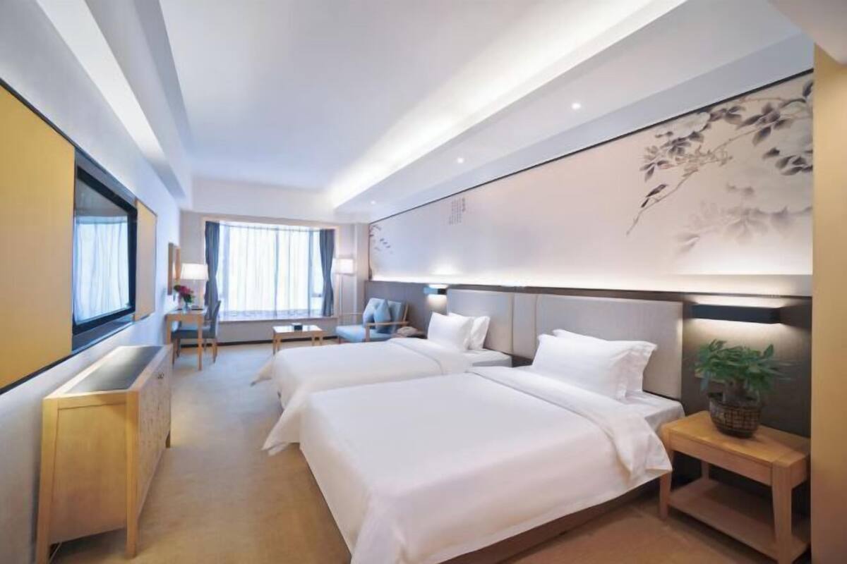 A hotel room with two beds, a desk, a chair, a window with curtains, and a wall with a floral mural.