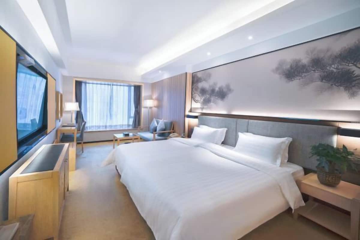 A modern hotel room with a large bed, a desk, a chair, a TV, and a large painting on the wall.
