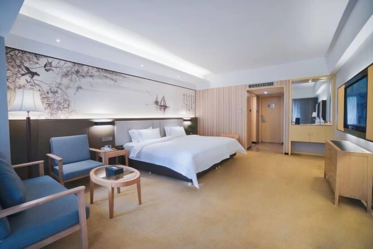 A modern hotel room with a large bed, a seating area with a sofa and armchairs, a small round table, and a large wall-mounted TV.