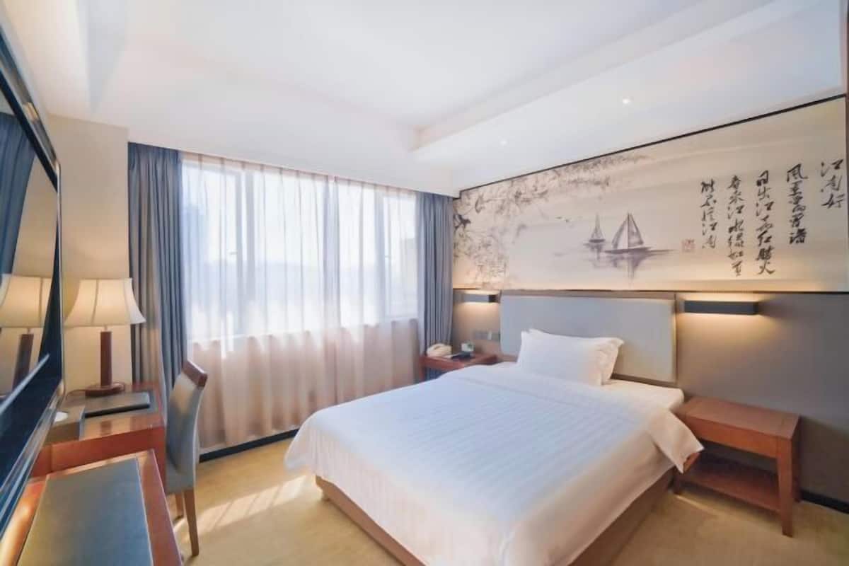 A hotel room with a large bed, bedside tables, a desk, a chair, and a large window with curtains.
