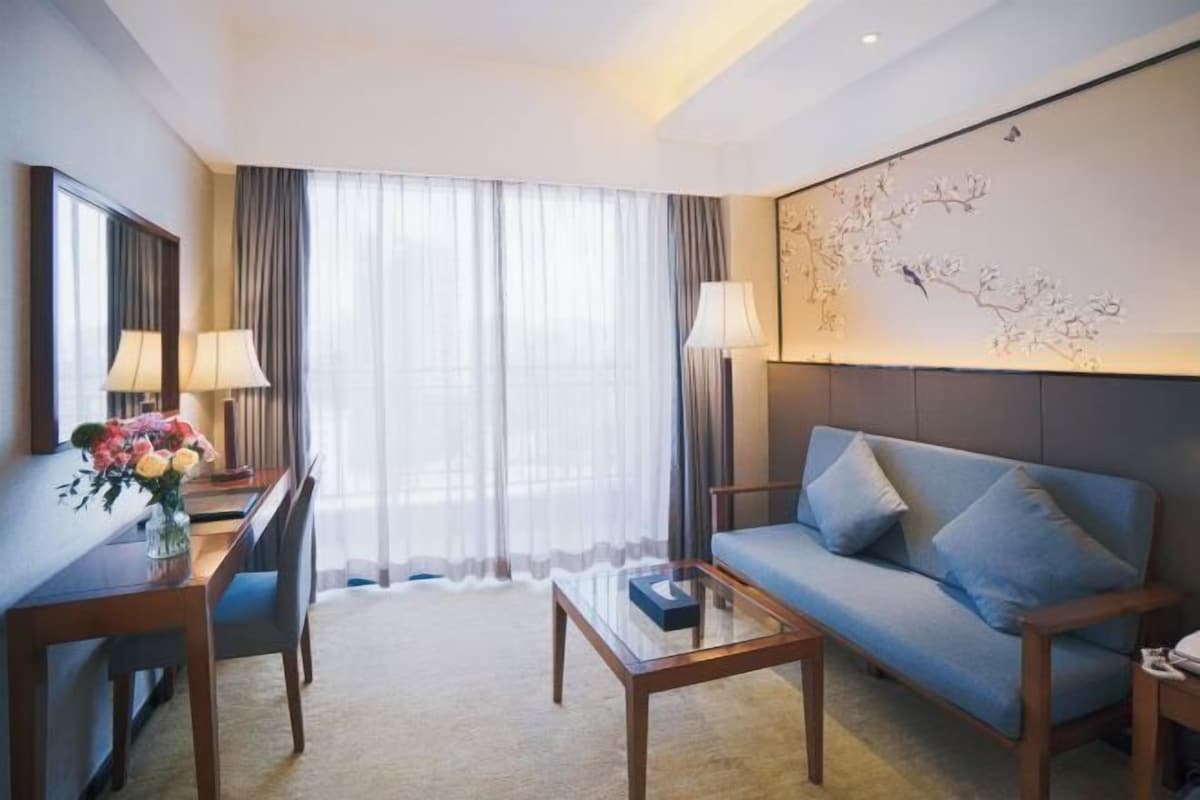 A modern hotel room with a sofa, a glass coffee table, a desk with lamps, and a large window with sheer curtains.