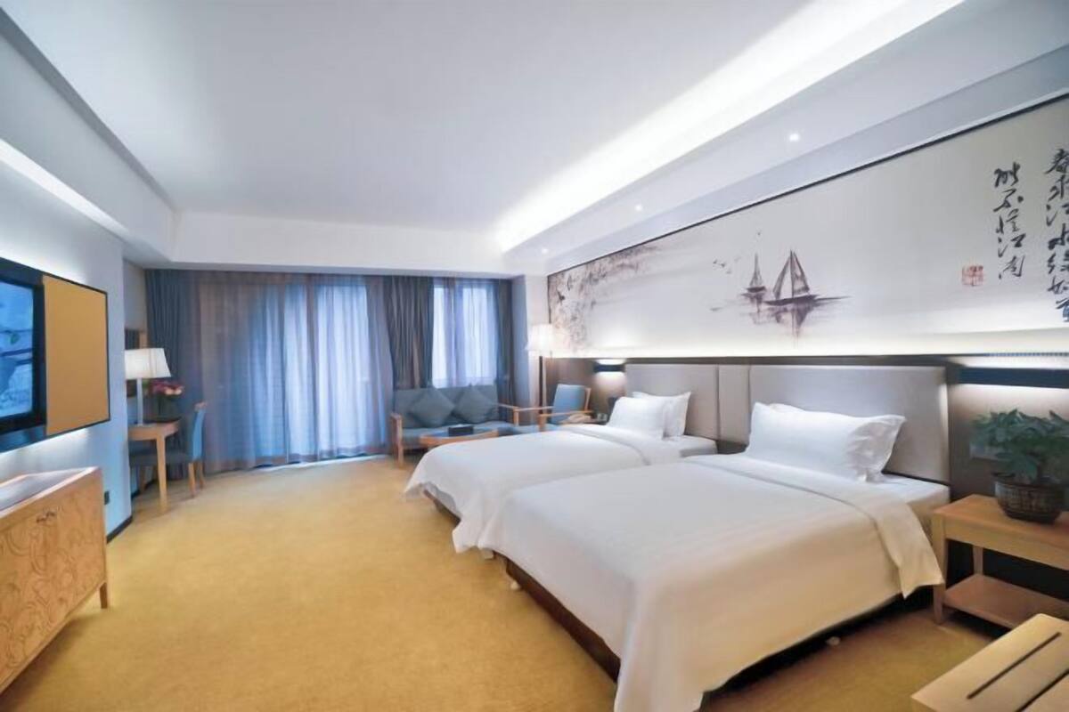 A hotel room with two beds, a desk, a chair, a TV, and a large artwork on the wall.