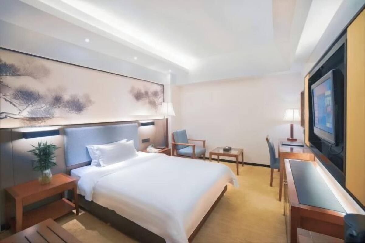 A hotel room with a bed, bedside tables, a desk, a chair, a television, and artwork on the wall.