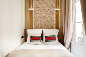 Una cama bien hecha con dos almohadas, un papel tapiz estampado y una ventana con cortinas.