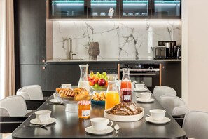 A breakfast setting with a table set for two, featuring croissants, fruit, juice, and a bread basket.