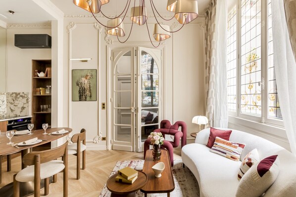 A well-lit living room with a dining area, a sofa with decorative pillows, and a large window with curtains.
