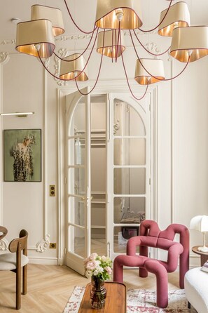 A room with a modern chandelier, a pink chair, a wooden coffee table with a vase, and a white bed.