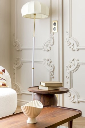 A room with a white ornate wall, a round wooden table with a white bowl, a tall floor lamp, and a white chair.