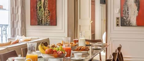 A breakfast table with fruits, pastries, and beverages in a well-lit room with artwork on the walls.