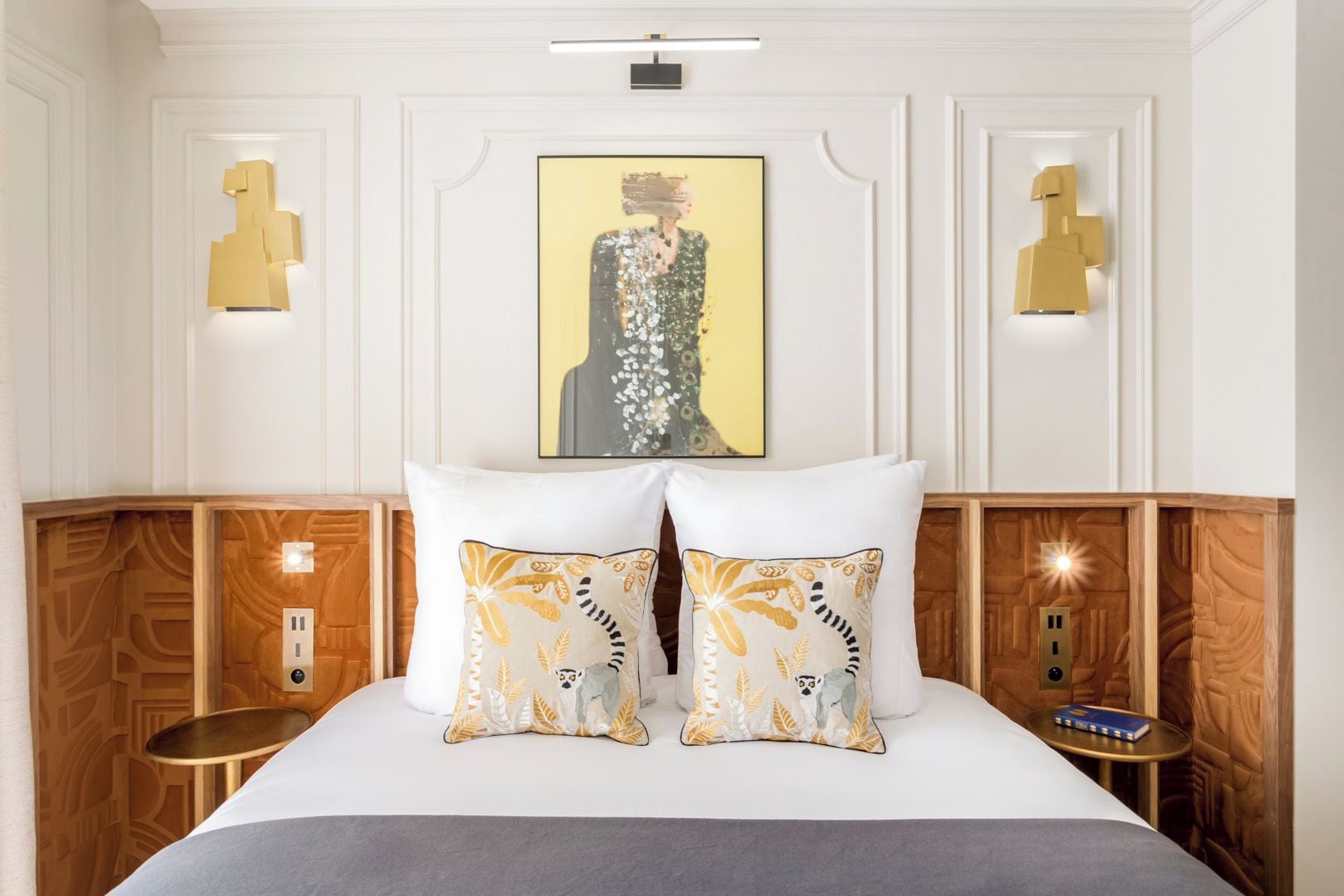 A hotel room with a neatly made bed, two decorative pillows, a framed artwork above the bed, and wall-mounted lighting fixtures.