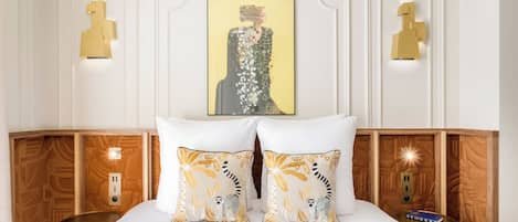 A hotel room with a neatly made bed, two decorative pillows, a framed artwork above the bed, and wall-mounted lighting fixtures.