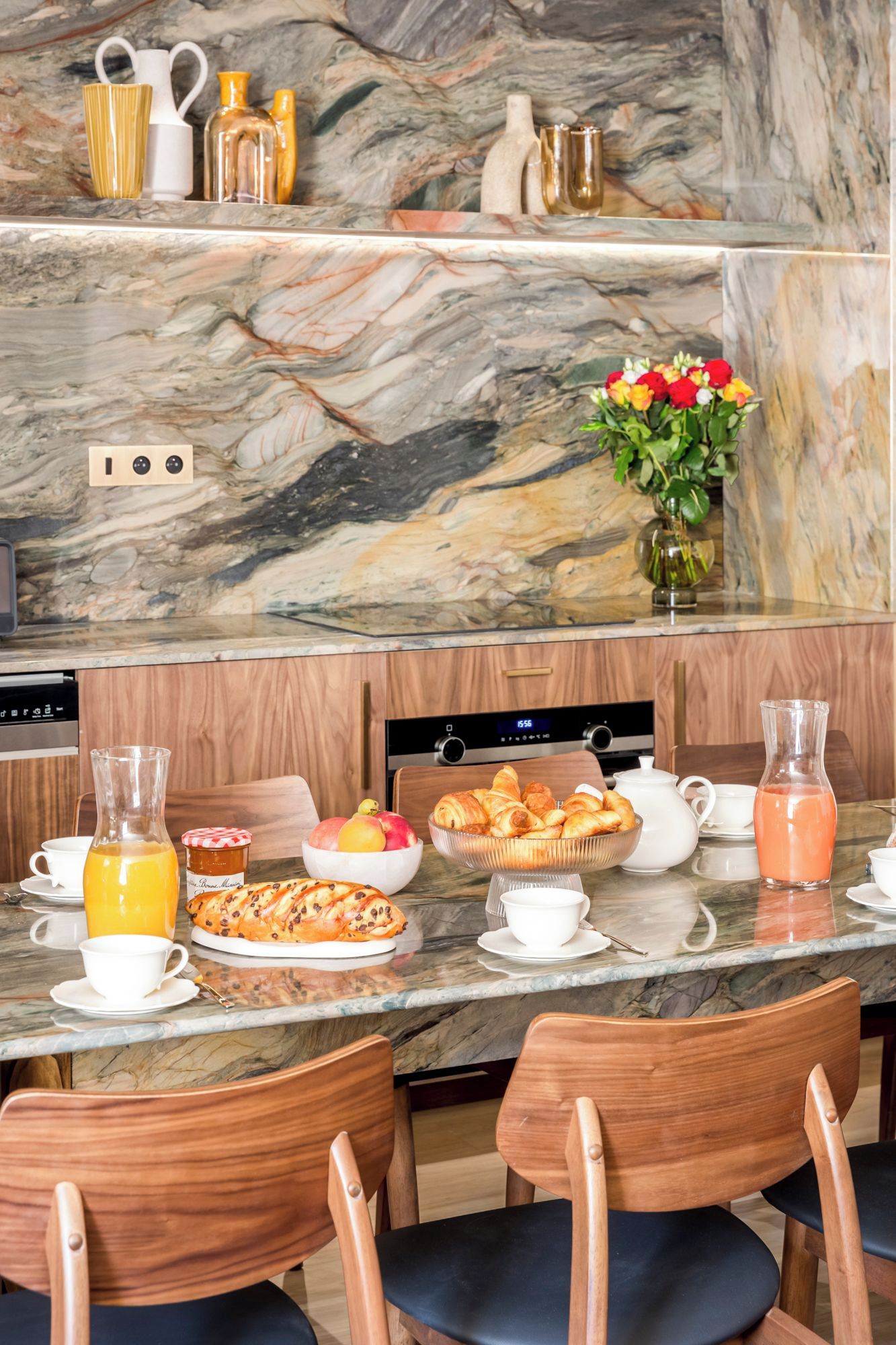 A breakfast setup with a marble countertop, wooden chairs, and a variety of breakfast items including pastries, fruit, and beverages.