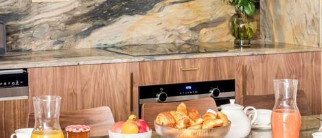 A breakfast setup with a marble countertop, wooden chairs, and a variety of breakfast items including pastries, fruit, and beverages.