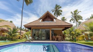 A resort with a swimming pool, palm trees, and a building with a triangular roof. - Rustic bungalow-beachfront condominium-017 (Tamandaré)