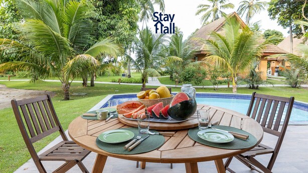 A dining table set outdoors with a pool in the background, surrounded by palm trees. - Rustic bungalow-beachfront condominium-017 (Tamandaré)