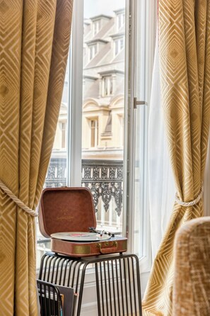 Ein Zimmer mit einem Vintage-Plattenspieler auf einem kleinen Tisch, Vorhängen mit geometrischem Muster und Blick auf Gebäude durch ein Fenster.