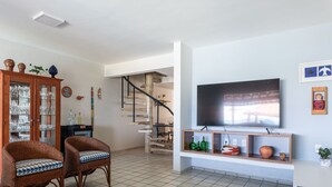 A living room with a TV, wicker chairs, a glass cabinet, and a staircase. - 4 Bedroom House on the Seaside of Carneiros with Private Pool and Cooking Service (Tamandaré)