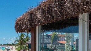 A beachside building with a thatched roof, large windows, and a view of the beach and palm trees. - 4 Bedroom House on the Seaside of Carneiros with Private Pool and Cooking Service (Tamandaré)