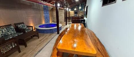 Un espacio interior amplio con una larga mesa de madera, una bañera de hidromasaje con azulejos azules y una zona de estar con muebles de madera.