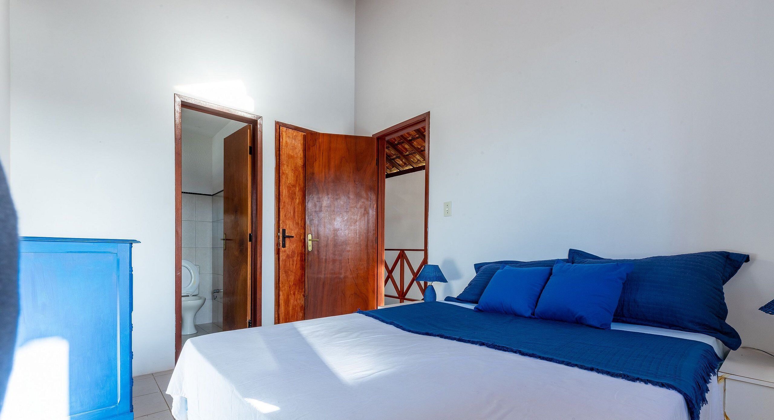 Un dormitorio con cama, un armario de madera, un baño visible a través de una puerta abierta y una lámpara azul.