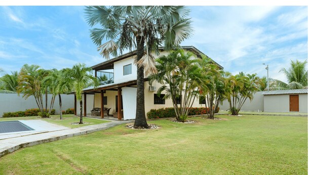 Une maison moderne à deux étages, dotée d’un patio couvert, entourée d’une pelouse bien entretenue et de palmiers.