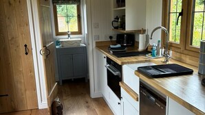 A compact kitchen with wooden flooring, a built-in oven, a dishwasher, and a window with a view of greenery. - Owls Nest - Alfriston East Sussex (Polegate)