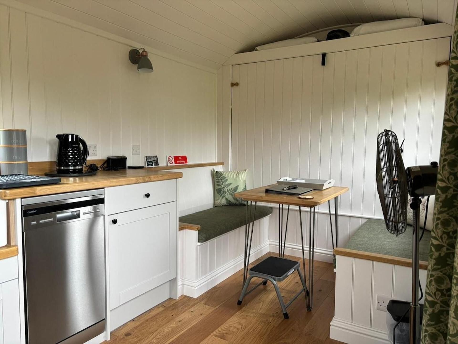 A compact kitchen with a dishwasher, a coffee maker, a small table, and a step stool.