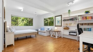 Ein Schlafzimmer mit einem Bett, einem Schlafsofa, einem Schreibtisch, einem Bücherregal und großen Fenstern mit Blick auf Grünflächen.