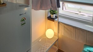 A compact room with a white mini-fridge, a hanging clothes rack with a checkered shirt, a potted plant, and a window with blinds. - Yeon Stay (Seoul)