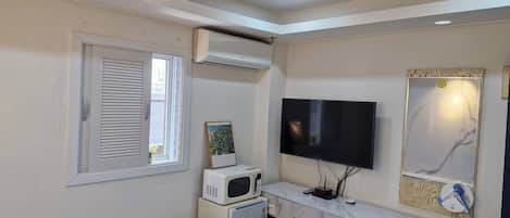 A bedroom with a bed, a desk with a computer, a TV, and a window with blinds.