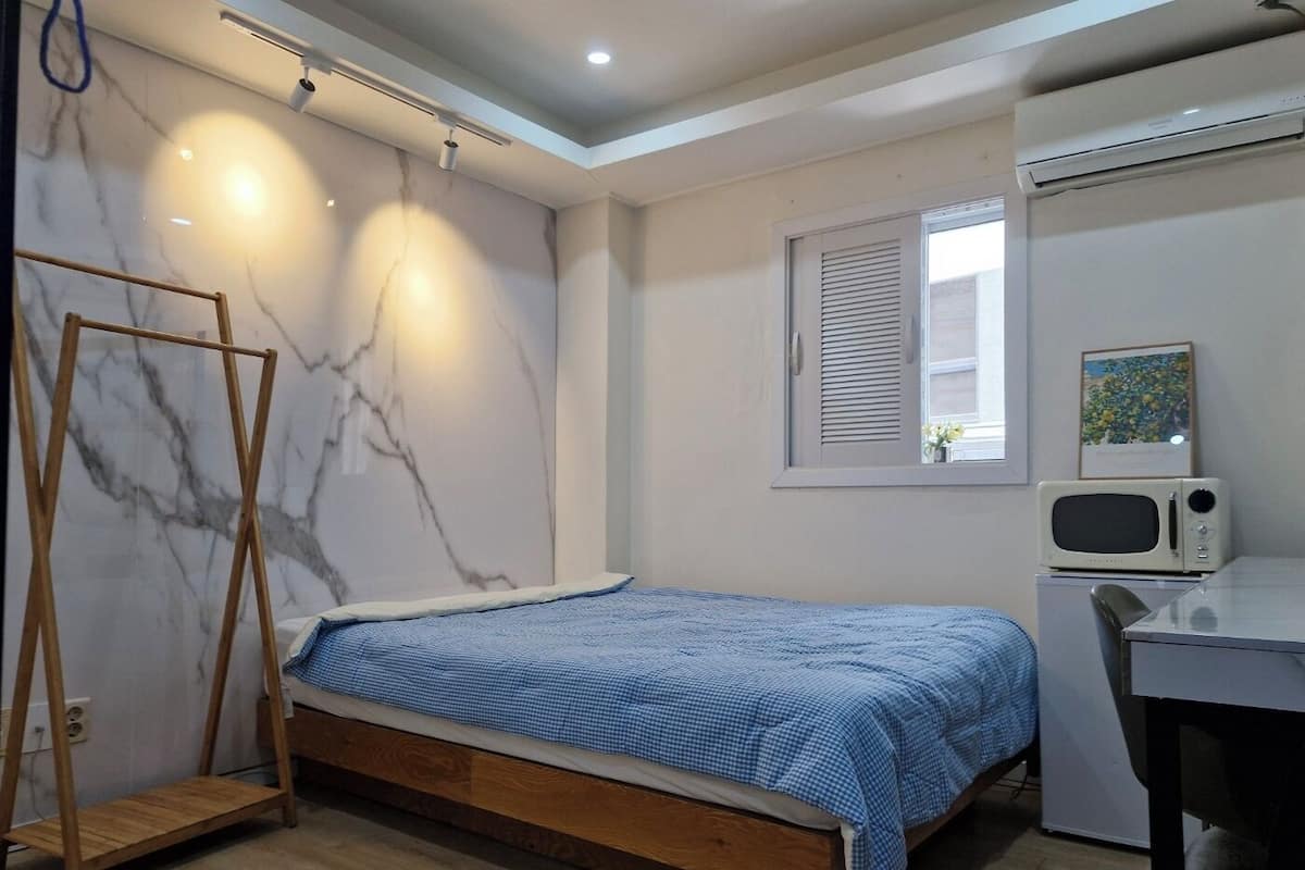 Un dormitorio con cama, una escalera de madera, un escritorio, una silla, un microondas y una ventana con persianas.